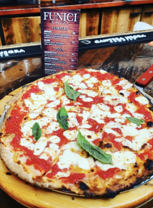 A thin-crust pizza topped with tomato sauce, melted mozzarella, and fresh basil leaves on a wooden board. A restaurant menu is visible in the background on a wooden bar counter.