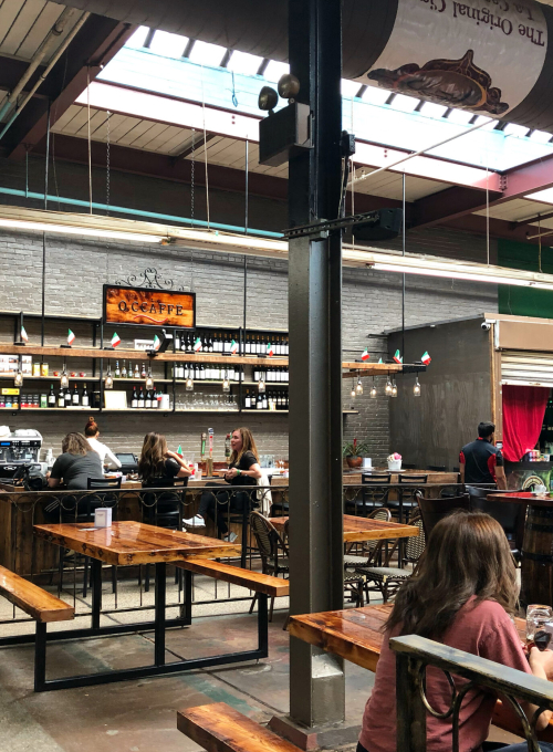 People dine at wooden tables inside a bright, spacious food hall with high ceilings. The counter and shelves behind display bottles and snacks, while festive bunting decorates the area. A family sits in the foreground eating.