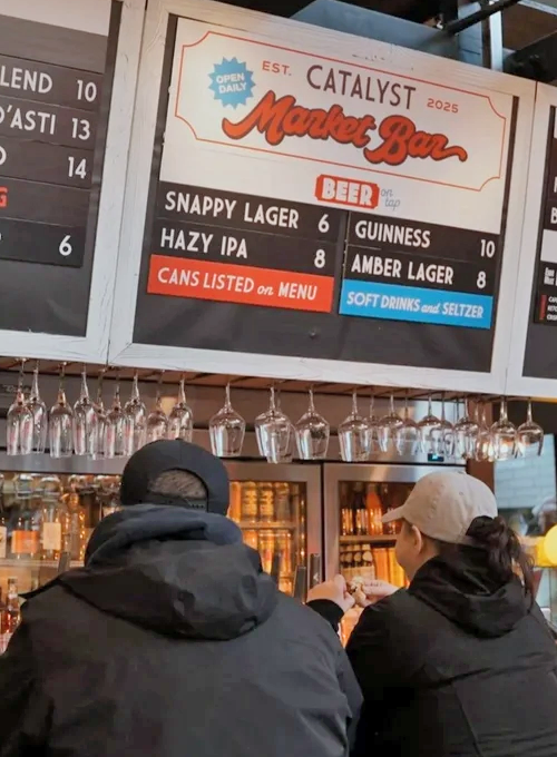 Two people in jackets sit at a bar counter. Above them, a Catalyst Market Bar menu lists beer options like Snappy Lager, Hazy IPA, Guinness, and Amber Lager, with prices shown. Glasses hang overhead.