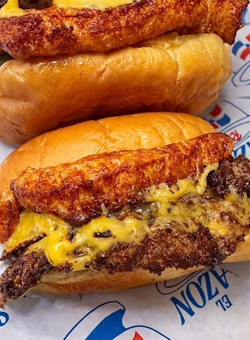 Two cheeseburgers on buns, each with a crispy fried cheese crust over melted cheddar and a beef patty, resting on branded paper.