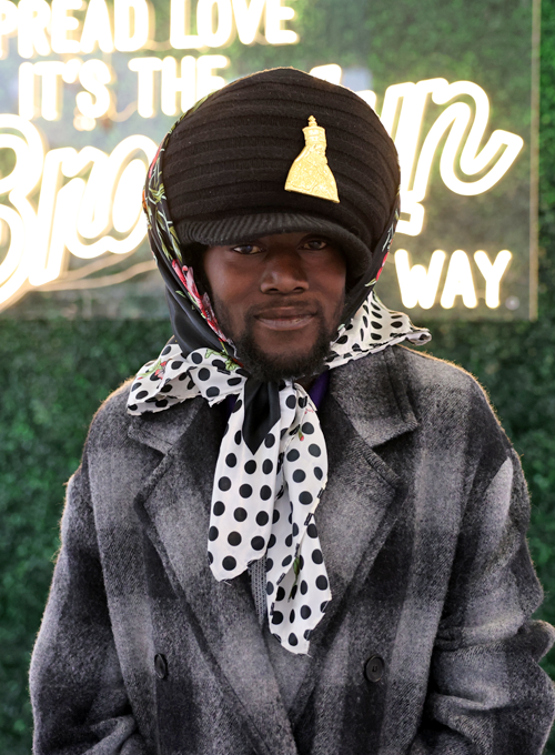 A person in a plaid coat, polka dot scarf, and black hat with a gold embellishment stands in front of a neon sign on a green leafy wall.