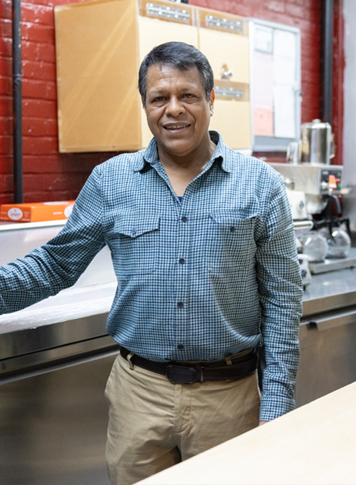 A man in a checked shirt smiles while standing in a kitchen with stainless steel counters, wooden cabinets, and a red brick wall in the background.