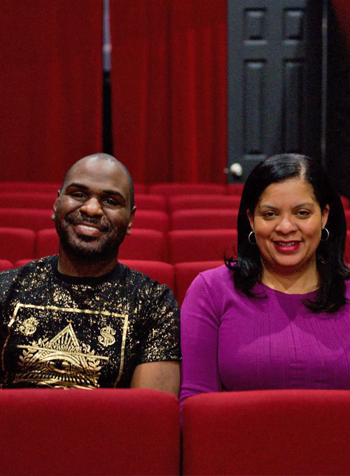 Two people are sitting side by side in red theater seats, smiling at the camera. Red curtains and a dark door are visible in the background.