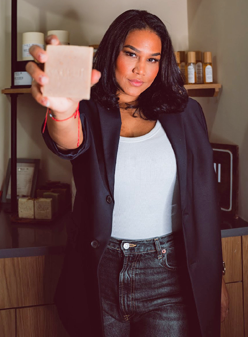 A woman with shoulder-length dark hair holds a bar of soap toward the camera, standing in a shop with wooden shelves lined with skincare products.