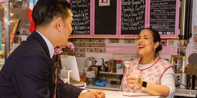A man in a suit leans on a counter, smiling at a woman in a pink apron holding a dessert behind the counter. Behind her, a chalkboard menu is visible. The setting is colorful and lively, with various decorations and small framed pictures on the wall. The shop is located in La Marqueta market. Photo by Kreg Holt.