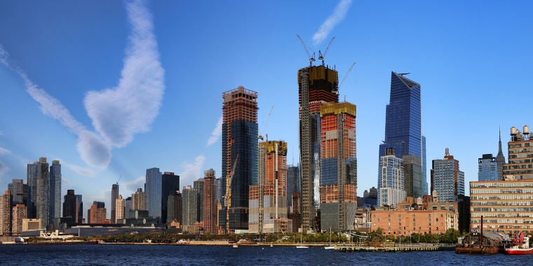 Panoramic view of the New York City skyline featuring modern skyscrapers of Hudson Yards under construction against a clear blue sky. A river is in the foreground, and fluffy white clouds add to the scenic urban landscape.