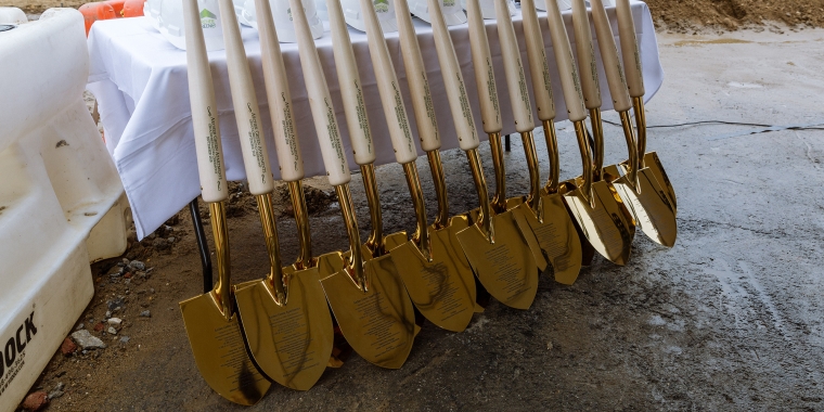 A row of gold-colored ceremonial shovels from the 168th Street ground breaking with light handles is lined up against a table covered with a white cloth. Hard hats are placed on the table, and construction materials are visible in the background.