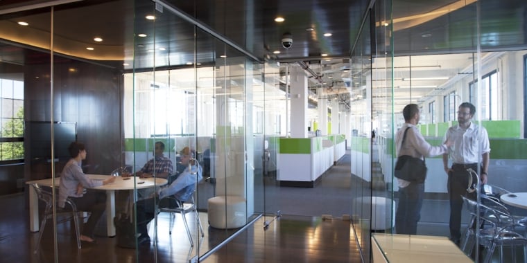 Modern office with glass walls and partitions. Employees are sitting at a round table and others are walking past outside. Green and white color scheme with large windows letting in natural light.