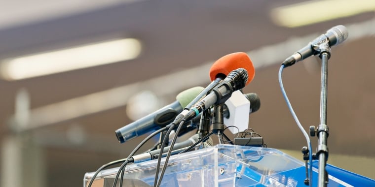 Multiple microphones in various colors are set up on a clear podium, ready for a speech or press conference. The background is blurred, focusing attention on the microphones.