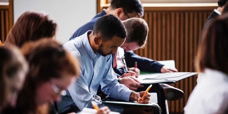 A group of LifeSci NYC applicants sitting at desks in a classroom, focused on writing in notebooks. The students are diverse, dressed in casual attire, and are positioned in rows. The setting is well-lit, with wooden paneling on the walls. Photo by Jordan Rathkopf/NYCEDC