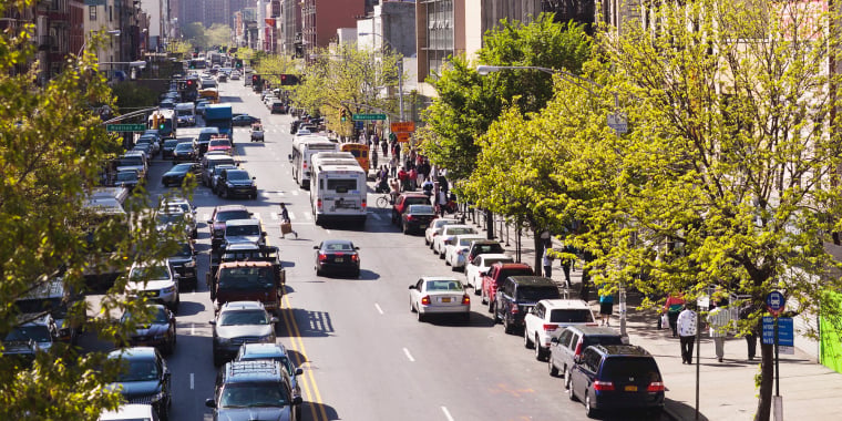 A busy 125th street in Harlem lined with trees, cars parked along both sides, traffic moving in both directions, and people walking on the sidewalks on a sunny day. Photo by Joe Buglewicz.