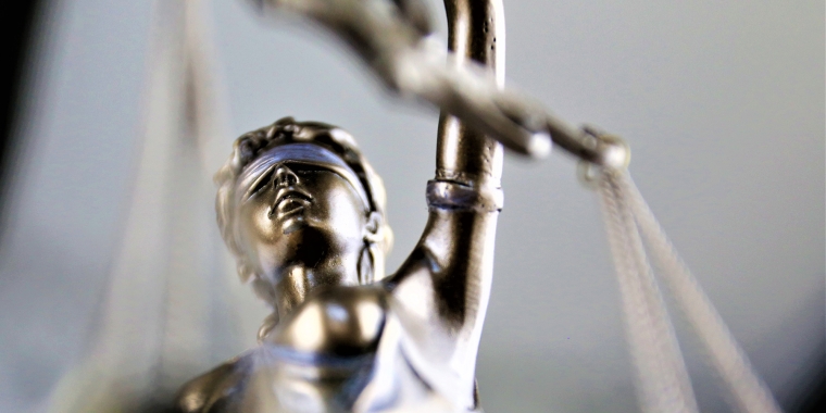 Close-up of a metallic statue of Lady Justice, featuring a blindfolded woman holding scales. The focus is on her face and raised arm, with the background softly blurred.