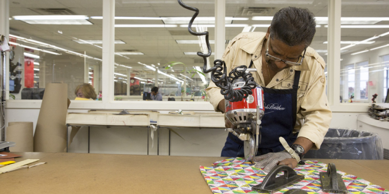 A fashion manufacturer using a circular fabric cutter on a colorful patterned cloth at a workbench in a sewing workshop. The room has large windows, and various sewing tools are visible on the table. Photo by Ian Spanier/NYCEDC.