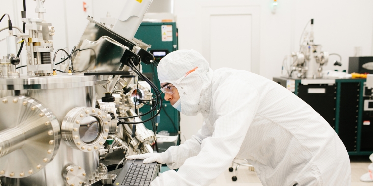 A scientist in a cleanroom suit operates a large, complex piece of laboratory equipment. They are bent over a computer keyboard, focusing intently. The room is bright and filled with high-tech machinery.