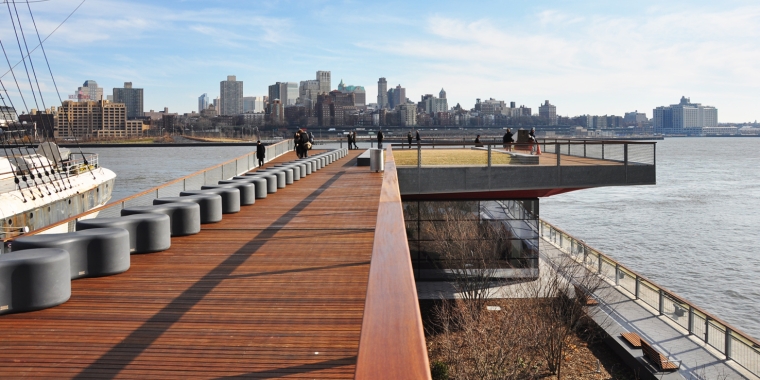 A waterfront boardwalk with modern seating extends toward a cityscape across the river. A large ship is partially visible on the left. People are scattered along the path, and the sky is bright with scattered clouds.