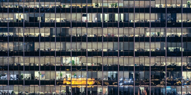 Night view of a high-rise office building with lit windows. Multiple floors are visible, showcasing office spaces with desks, computers, and scattered decor. The building reflects city lights and activity from within.
