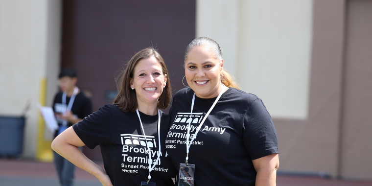 Two female NYCEDC employees at the BAT Block Party stand side by side outdoors, smiling at the camera. They are wearing matching black t-shirts and lanyards. A blurred background features a person walking and a building section.