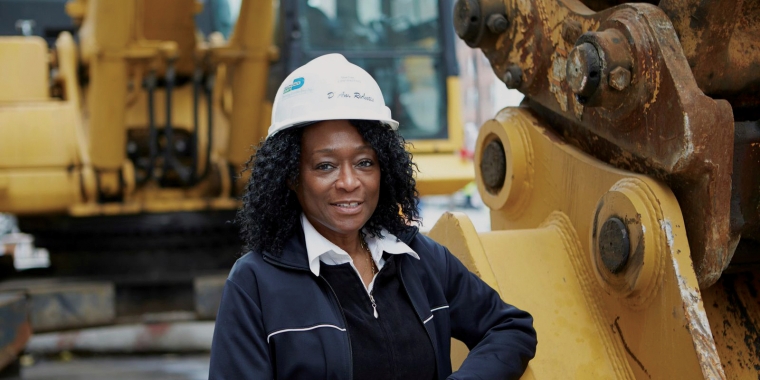 D&#039;Ann Ricketts, Owner of BarDen Contracting Services, LLC, wearing a white hard hat and dark jacket stands confidently next to large yellow construction machinery. She is outdoors, with more equipment visible in the background.
