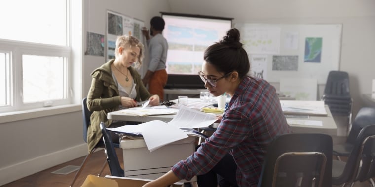 Two Not-for-Profit Bond Program working in a bright office. One is seated, looking at documents on a table. The other, wearing a plaid shirt and glasses, is seated and searching through a box. A third person, blurred, stands at a screen in the background.