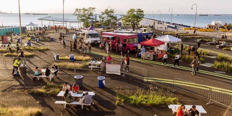 Aerial view of BAT Block Party, showing a waterfront food festival featuring food trucks, vendor tents, and picnic tables. People are enjoying food and mingling. The scene is set against a backdrop of water and a clear sky, with ships visible in the distance. Photo by Kreg Holt/NYCEDC.