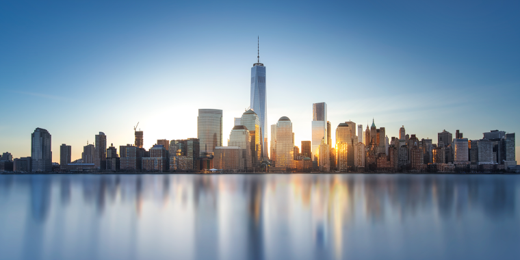 A panoramic view of NYC skyline at sunrise, with tall skyscrapers reflecting on the calm water in the foreground. The sky is clear and blue, and the sunlight illuminates the buildings, creating a serene and picturesque scene.