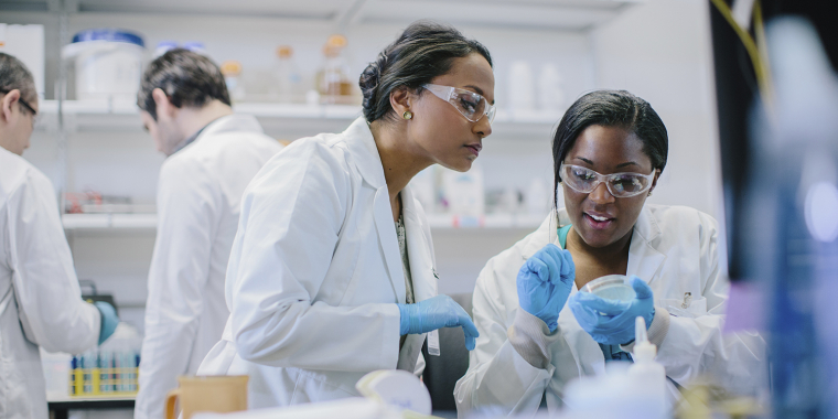 Two scientists in a lab are wearing white coats, gloves, and safety goggles. They are discussing data. Another scientist works in the background. The lab is filled with equipment and supplies.