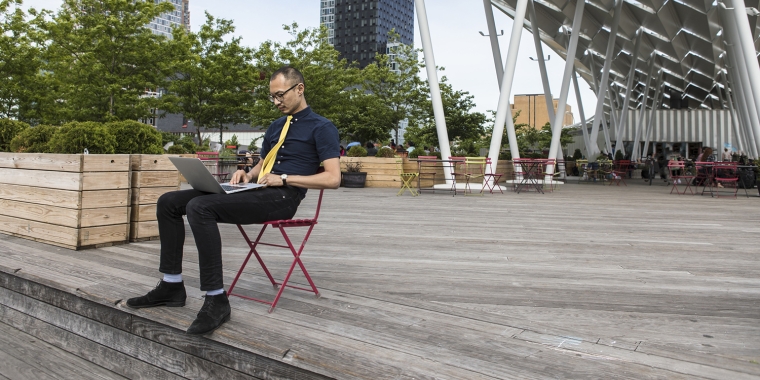 Show thumbnail preview A person sitting on a red chair on a wide wooden deck under a large modern canopy structure, working on a laptop. The background features city buildings and green trees, with a partially cloudy sky.
