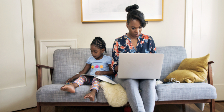 A Black woman sits on a gray couch working on a laptop while her young child beside her uses a tablet. They are both focused on their devices in a cozy, well-lit living room with pillows and a rug.