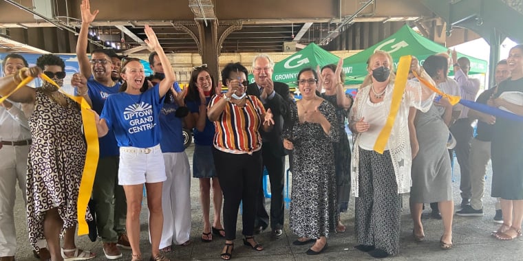 A diverse group of people cheer and smile while holding and cutting a long yellow ribbon under an outdoor structure at the 125 Pedestrian Plaza ribbon-cutting ceremony. Some hold scissors and wear event t-shirts or casual summer clothing.