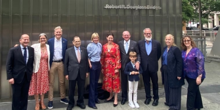 A group of ten adults and one child pose together, smiling, in front of a building with the sign “Robert R. Douglass Bridge” visible above them. Trees and urban structures are in the background.