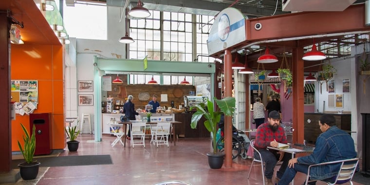 Indoor view of La Marqueta Market with a few people seated at tables. The space has high ceilings, red and orange walls, and greenery. There are various food stalls, and a large window allows natural light to fill the area.