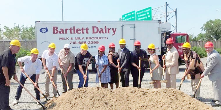 A group of people in hard hats and business attire holding shovels and posing at a groundbreaking site with a mound of dirt in front of a Bartlett Dairy truck.