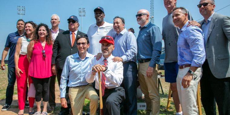 A group of men and women, some in business attire and others casually dressed, pose together in Staten Island Ballpark under a sunny sky. One man in front wears a red cap and holds a baseball bat.