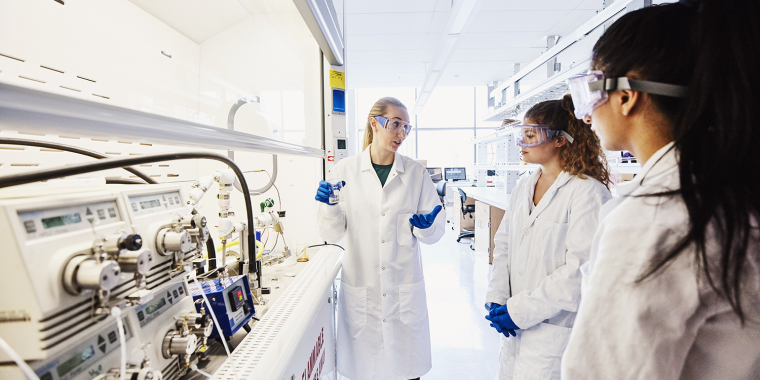 Three scientists wearing lab coats and safety goggles discuss in a bright laboratory next to scientific equipment and machinery. One person gestures while holding a tablet, and the others listen attentively.