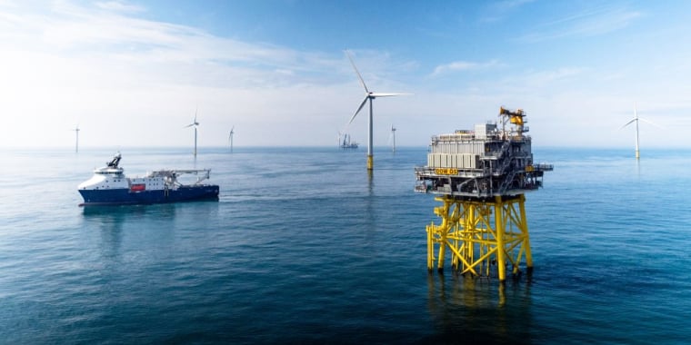 Large offshore wind platform with yellow supports stands in the sea near a service ship. Wind turbines are visible in the distance under a blue sky with light clouds.