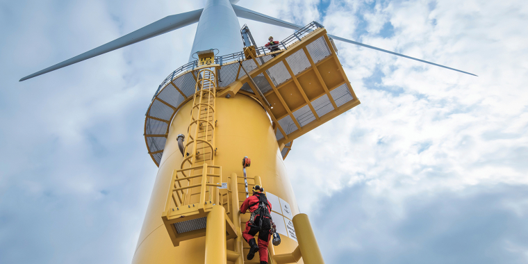 Worker in red safety gear climbs a yellow ladder on a large offshore wind turbine tower. Another person is visible on a platform above. The sky is cloudy, and the turbine blades extend outward from the top.