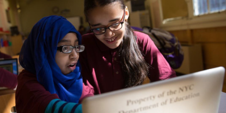 Two female students at P.S. 188, The Island School, one wearing a blue hijab, look excitedly at a laptop with Property of the NYC Department of Education on it. They're in a classroom with sunlight streaming through the windows.