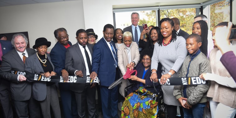 A diverse group of people stands together indoors for CACCI Headquarters ribbon cutting ceremony. A woman in a wheelchair cuts the ribbon with large scissors while others smile and gather around, holding the ribbon labeled NYCEDC.