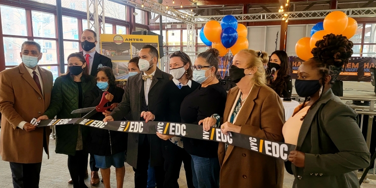 Group of people wearing masks stand indoors, holding and cutting an NYCEDC ribbon at La Placita Reopening. Orange and blue balloons are in the background, along with banners and signs.