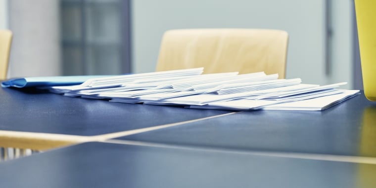 A stack of white envelopes is spread across a dark blue tabletop. Wooden chairs are partially visible in the background. The scene suggests a setting for organizing or distributing paperwork.