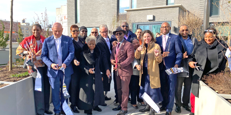 A diverse group of people in formal attire, smiling and holding ribbon-cutting scissors, stand outside Archer Green in southeast Queens to celebrate the opening of the new affordable housing development.