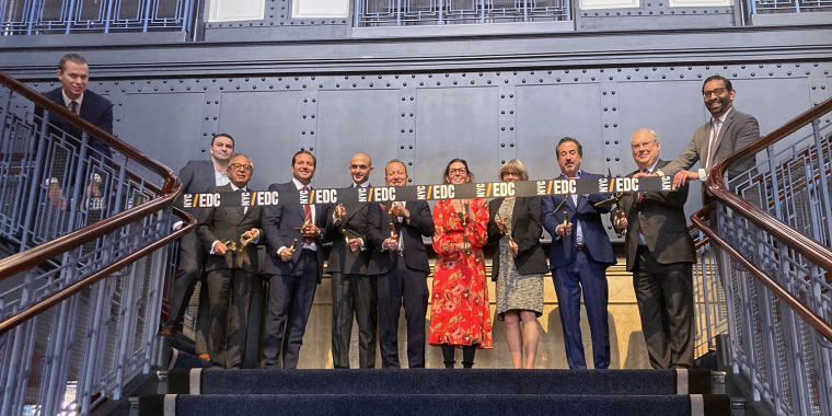 A group of eleven people stands on a staircase, holding trophies and a large ribbon with NYCEDC on it, smiling in front of a blue and gray wall for a photo at a ribbon cutting of the Battery Maritime Building.