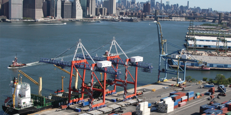 Busy port with colorful shipping containers, cranes loading a cargo ship, and tugboats, set against New York City skyline with tall buildings across the river under a clear sky.