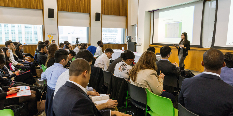 A woman stands in front of a screen giving a presentation to seated LifeSci interns in a modern, bright conference room with large windows. The interns are dressed in business attire and appear engaged.