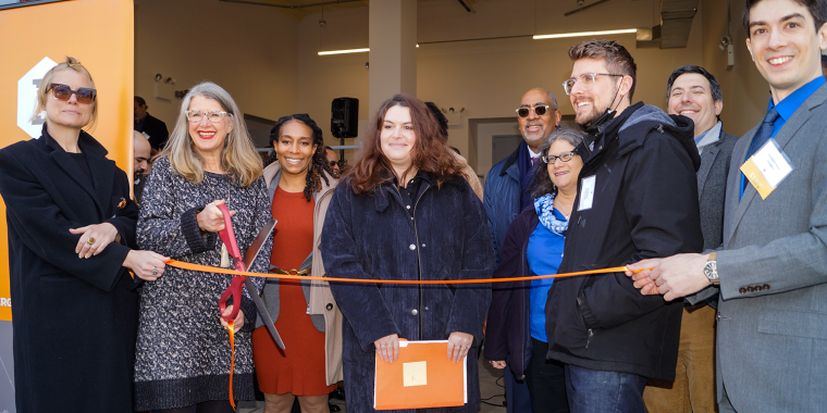 Group of people smile as a woman cuts an orange ribbon indoors at a 500 Stagg ribbon-cutting ceremony. Some hold the ribbon while others watch, posing for a celebratory photo.