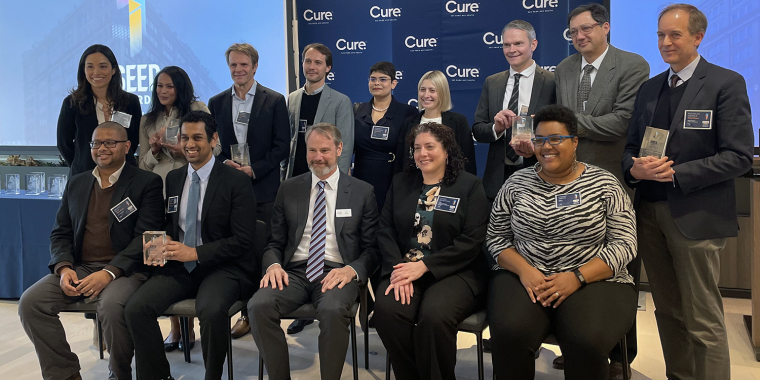 Group of fourteen XSeed Award winners, some seated and some standing, pose indoors while smiling and holding glass awards in front of a Cure banner and a screen displaying the word XSeed.