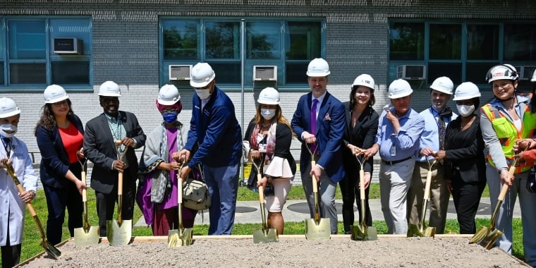 A group of people in formal and construction attire, wearing white hard hats, stand in a row outdoors, each holding a golden shovel and posing for a Metropolitan Floodwall groundbreaking ceremony in front of NYC Health + Hospitals/Metropolitan in East Harlem.