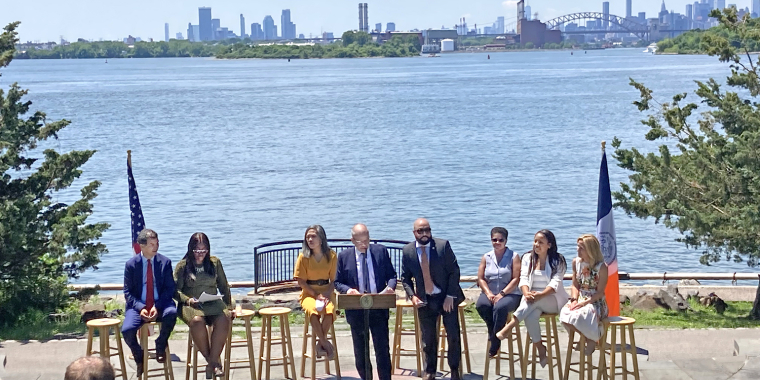 Group of eight community leaders sit on stools near a waterfront, with one person standing at a podium. Cameras film the release of Hunts Point Forward Vision, New York City skyline and bridge visible in the background, and two flags are displayed on each side.