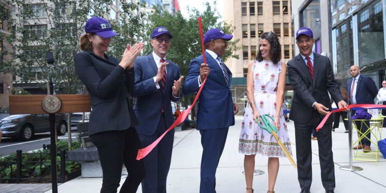 Group of five people in business attire and purple hats at Hudson Square Streetscape Ribbon Cutting in New York City. Two people hold giant scissors, while others clap and smile on the sidewalk. Mayor Eric Adams is in the middle.