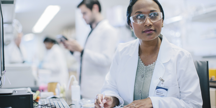 Female scientist wearing safety glasses and a lab coat sits at a lab desk, writing on a clipboard. Laboratory equipment and other scientists working in the background are visible.